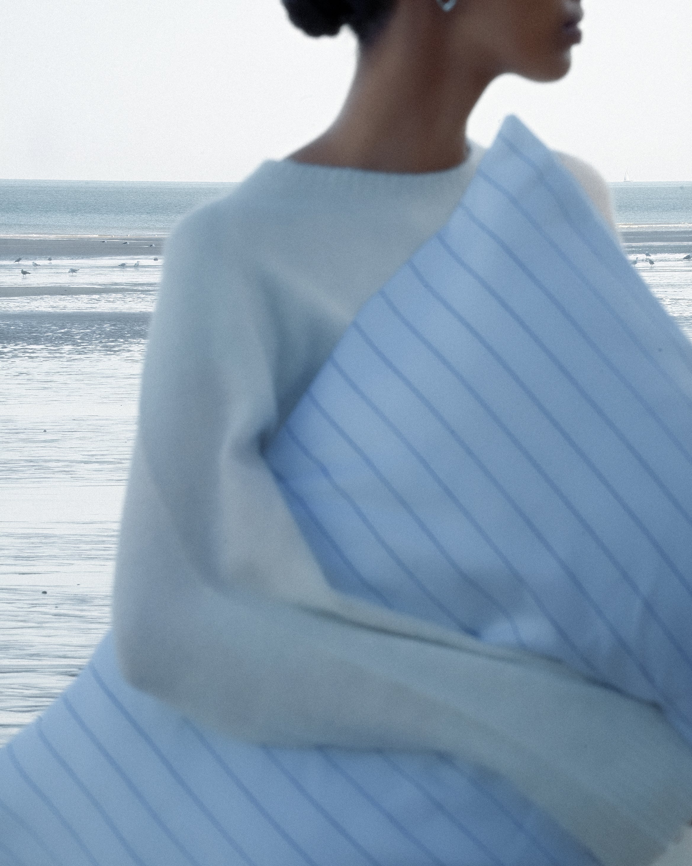 Blurry image of a woman carrying a pinstripe pillow, standing in front of the blue ocean.