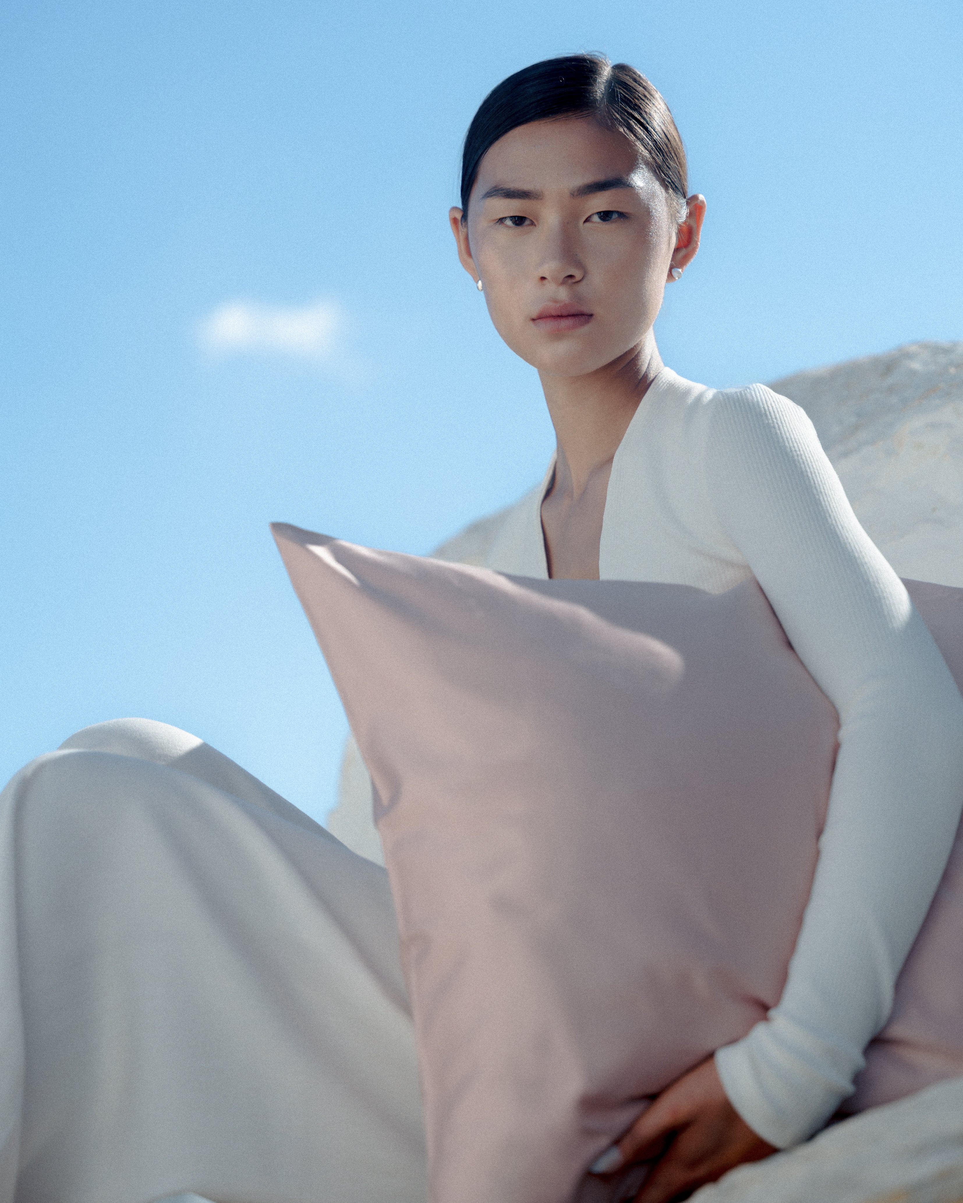 woman carrying a soft big light pink pillow, sitting on a rock in Mykonos