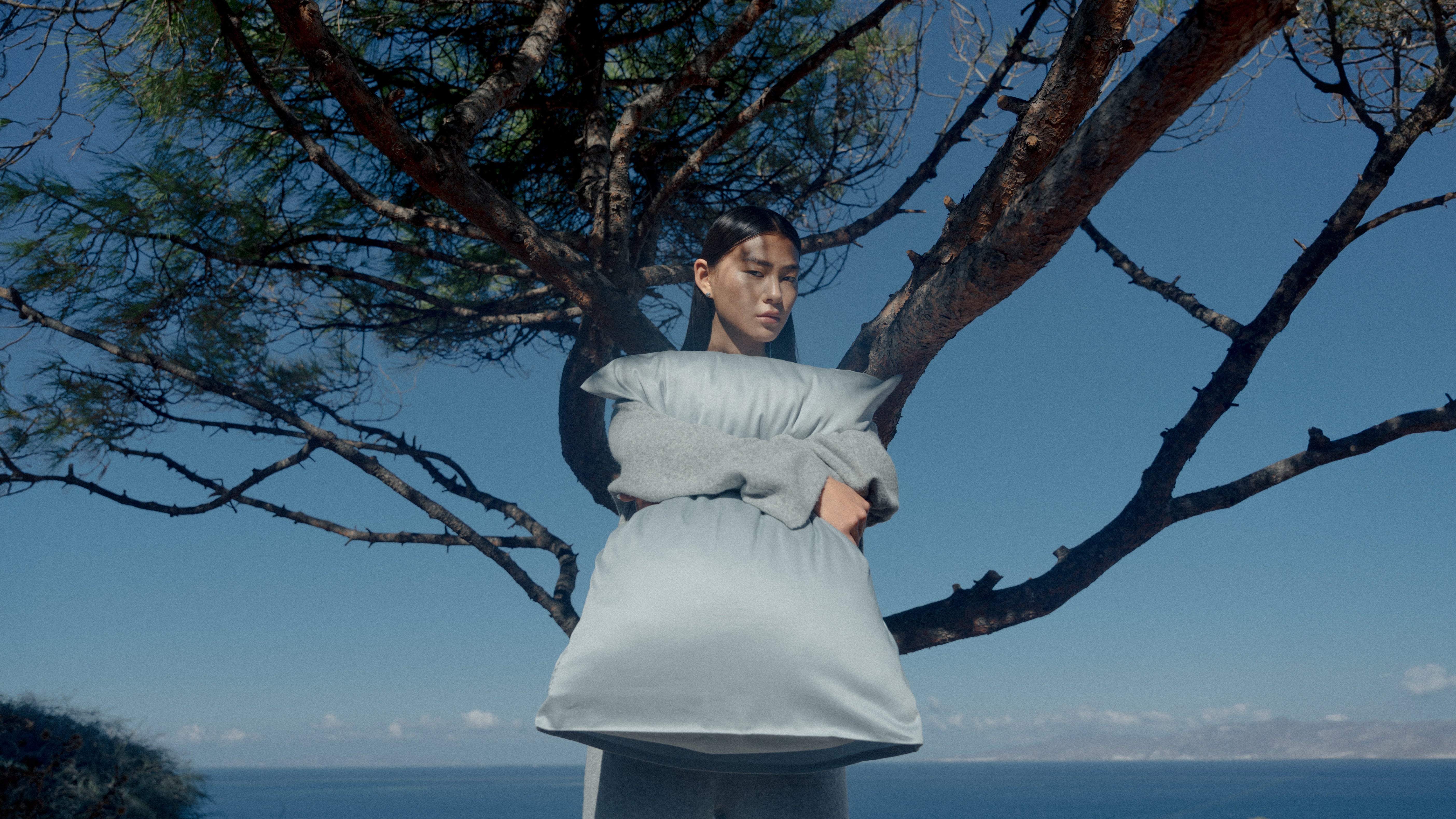 Kvinde med pude i armen, foran et smukt træ og med blå baggrund.